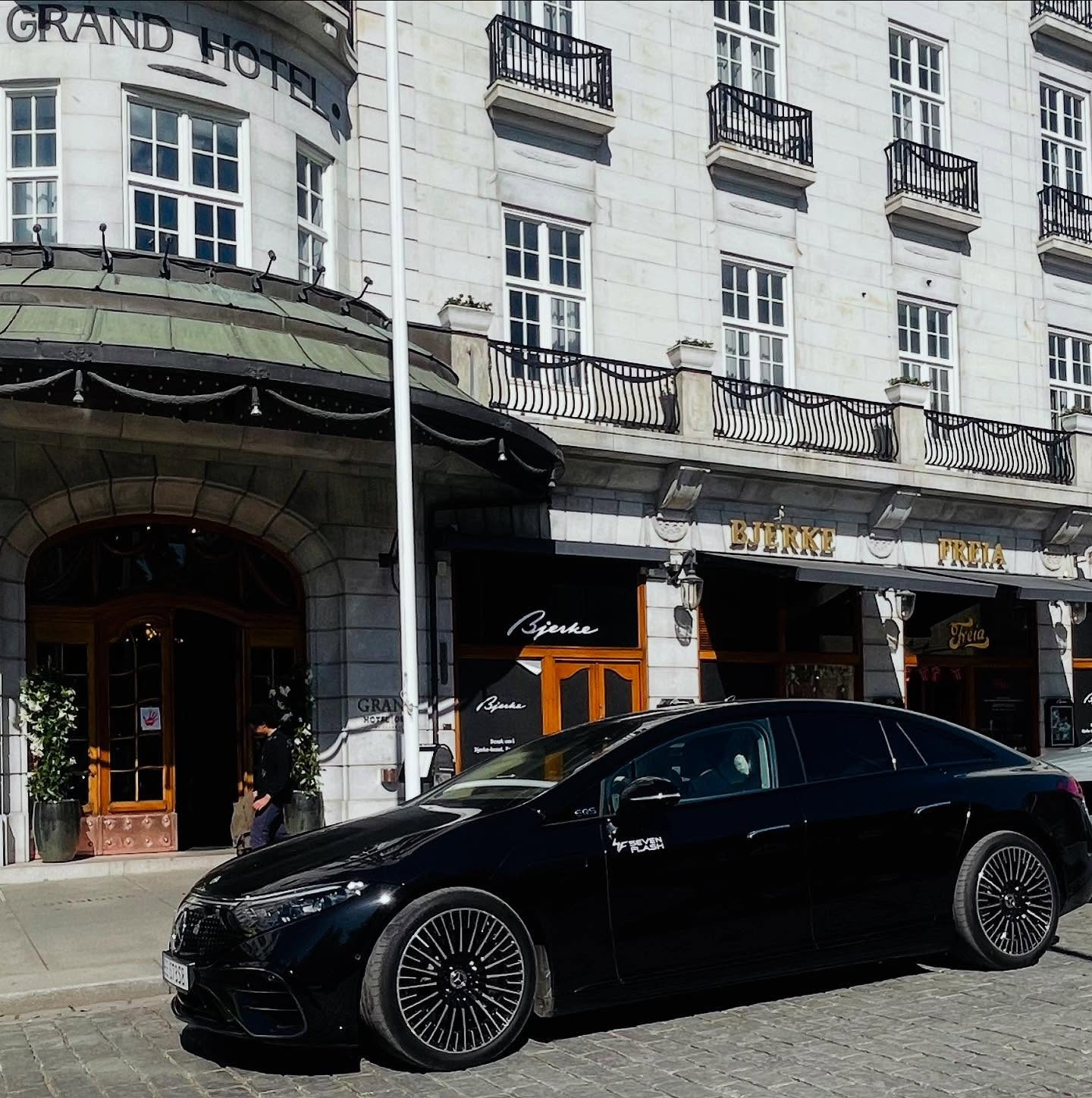 Black luxury Mercedes sedan parked outside Grand Hotel in Oslo, with the hotel’s classic white façade and storefronts in the background.